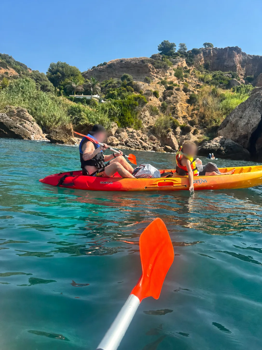 An adult and a young child in life jackets paddling a red and yellow sit-on-top kayak over clear turquoise water below the cliffs near Nerja