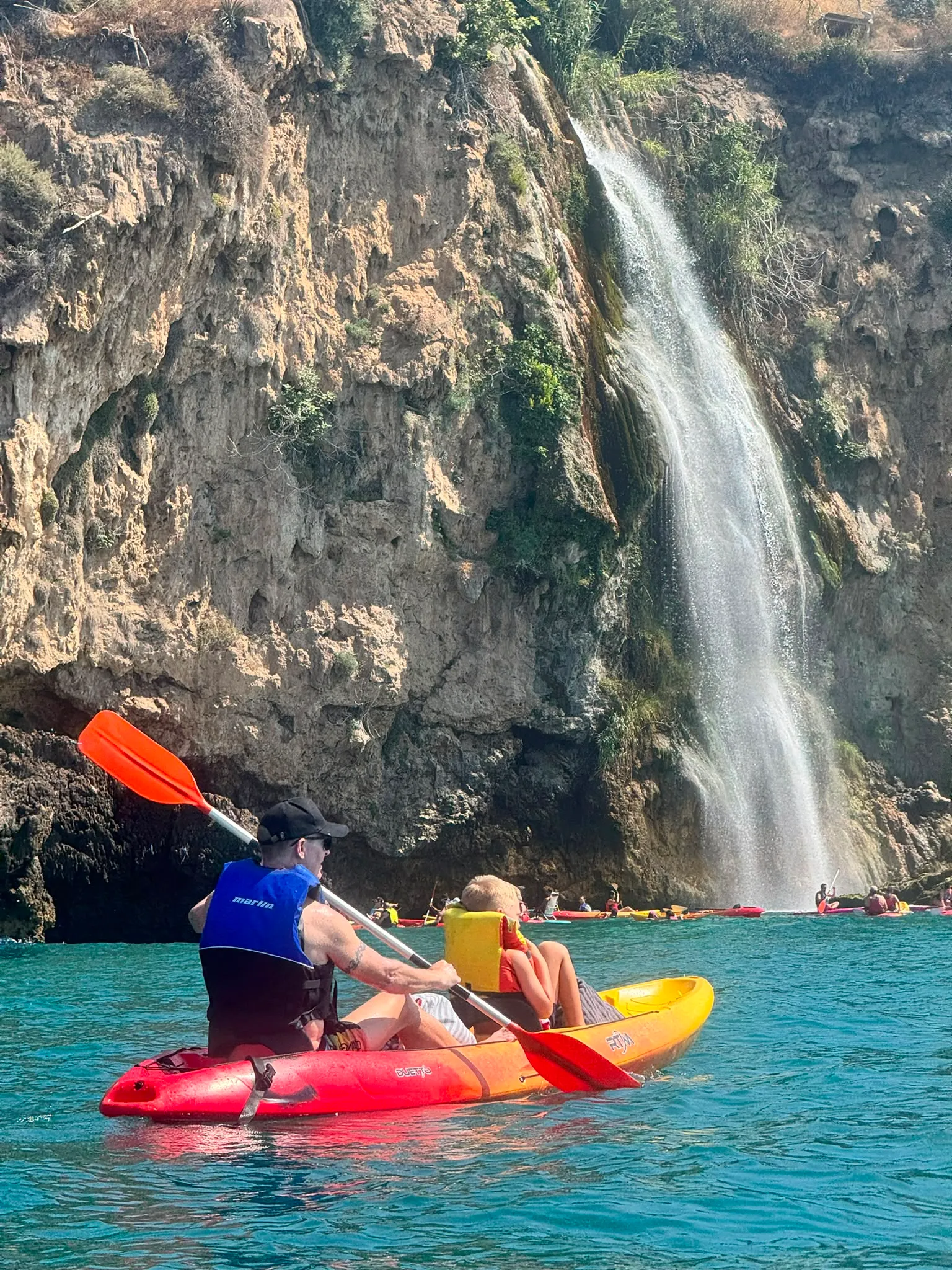 A kayak approaching the Maro waterfall, where water falls down a limestone cliff straight into the sea