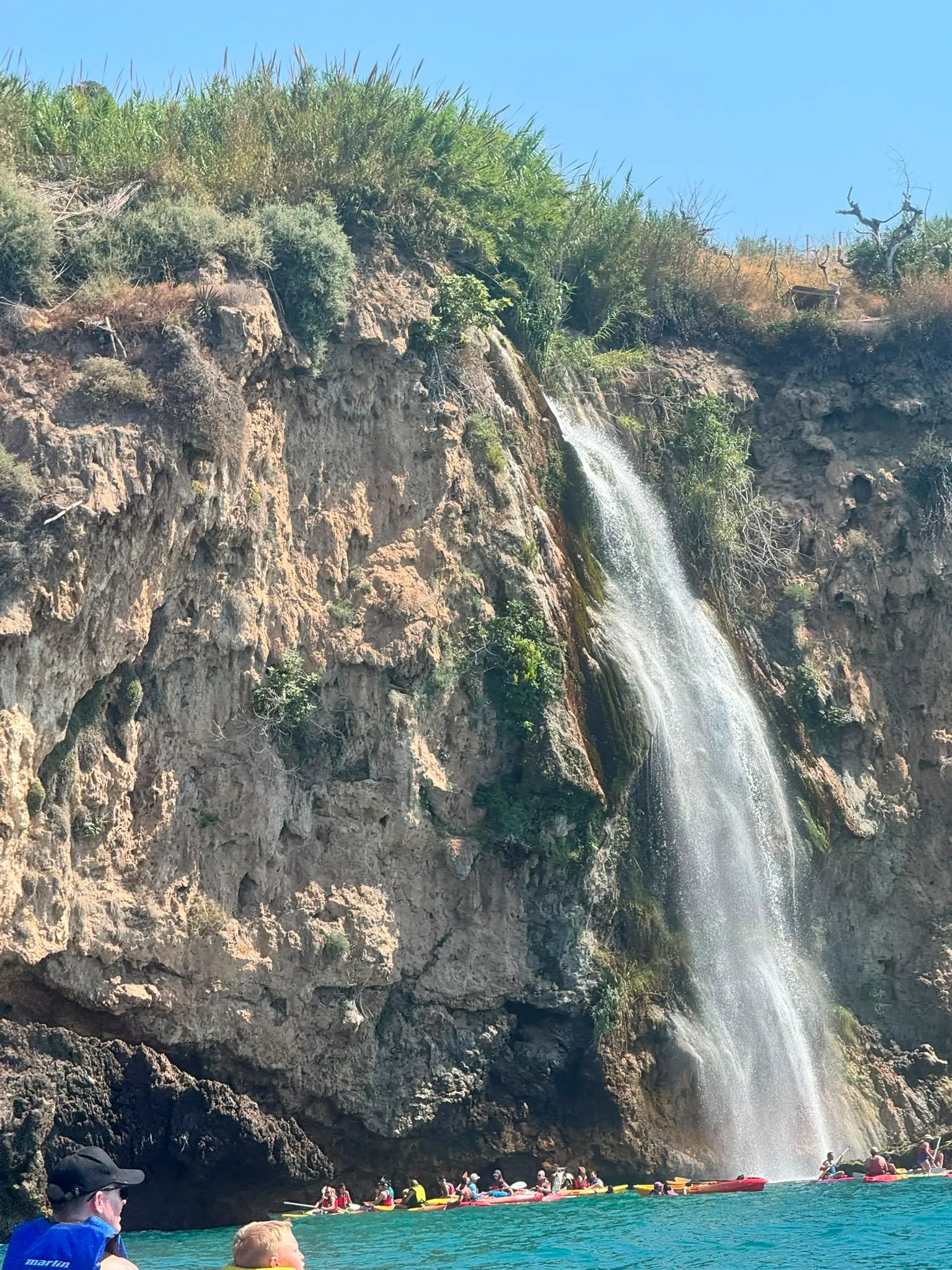 The Maro waterfall dropping down a tall limestone cliff into the sea near Nerja, with a line of kayaks resting at its base